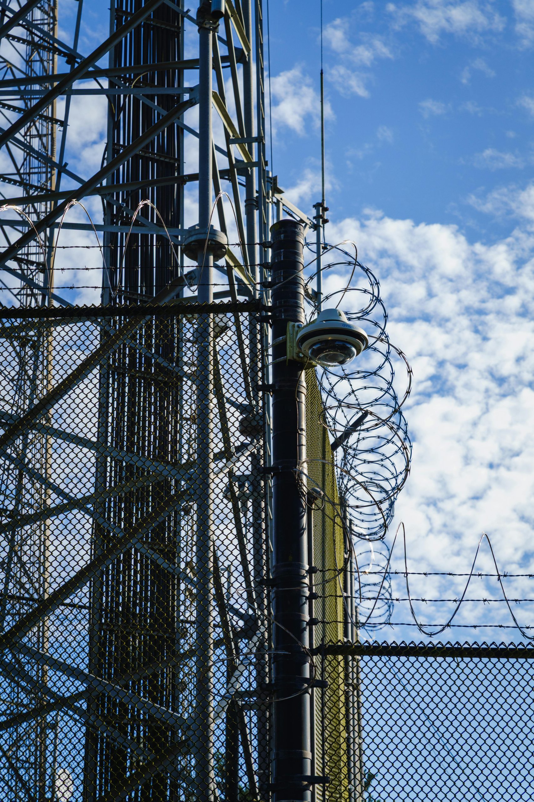 Cell tower structure featuring barbed wire and fencing under a bright blue sky with clouds.
